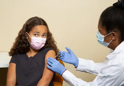 A masked doctor in a white coat and blue gloves administers a vaccine into the arm of a masked child. A masked doctor in a white coat and blue gloves administers a vaccine into the arm of a masked child.